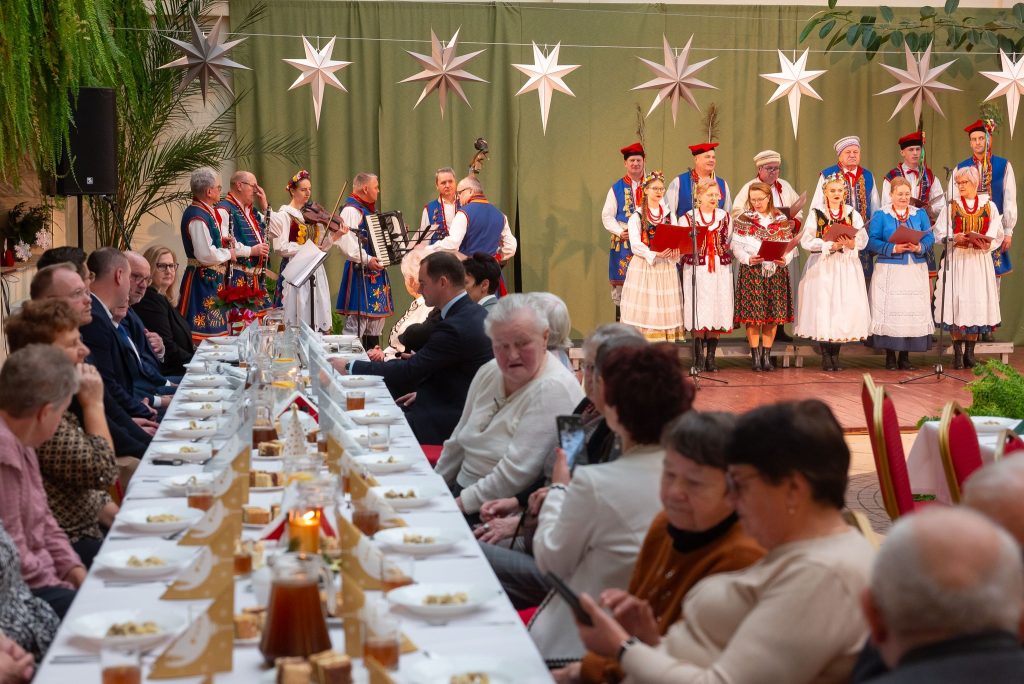 goście siedzą przy zastawionych stołach na atrium Centrum Kultury Wsi Polskiej w Wierzchosławicach a w tle widoczny śpiewający Zespół Pieśni i Tańca Swojacy w tradycyjnych strojac