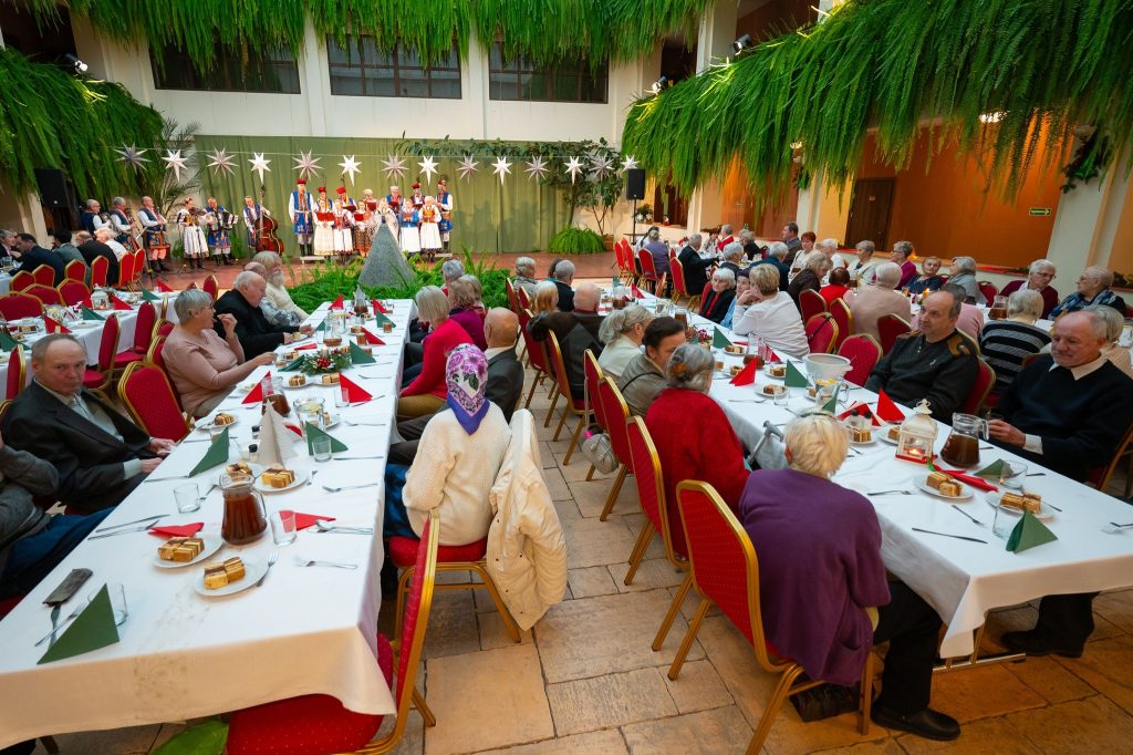 goście siedzą przy zastawionych stołach na atrium Centrum Kultury Wsi Polskiej w Wierzchosławicach a w tle widoczny śpiewający Zespół Pieśni i Tańca Swojacy w tradycyjnych strojach