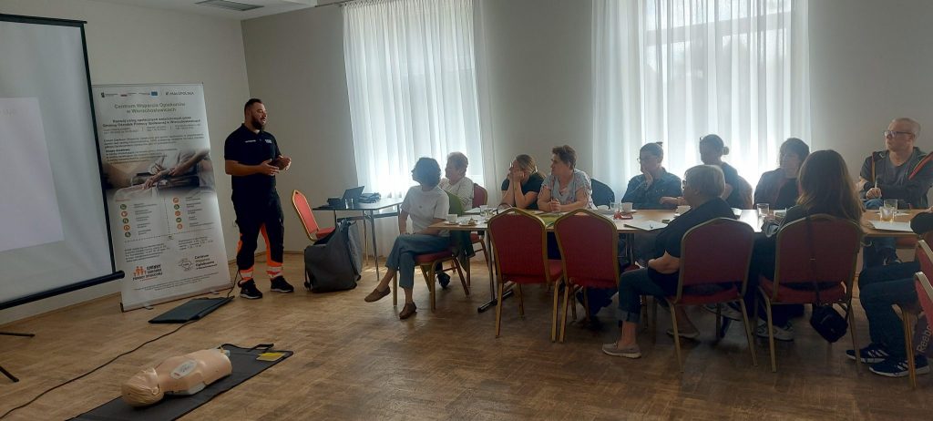 zdjęcie przedstawia grupę ludzi siedzących przy stole i słuchających wykładu instruktora